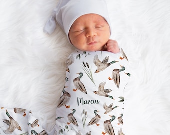 May include: A white swaddle blanket with a pattern of green reeds, brown and green ducks, and the name "Marcus" repeated. A baby is swaddled in the blanket and wearing a white knit hat.