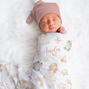 May include: A newborn baby wrapped in a white swaddle blanket and wearing a mauve knotted hat. The blanket is decorated with princess, frog, butterfly, and star illustrations. The name "Amelia Rose" is printed on the blanket.