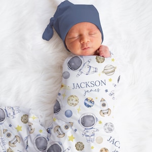 May include: A white swaddle blanket with a blue and yellow astronaut, rocket, and planet pattern. The blanket is wrapped around a baby wearing a blue knitted hat. The blanket has the name "JACKSON james" printed on it.