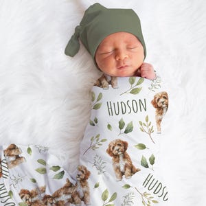 May include: A newborn baby wrapped in a white swaddle blanket and wearing a green hat. The blanket features a repeating pattern of brown puppies, green leaves, and the name "HUDSON" in green lettering. The baby is lying on a white, fluffy surface.