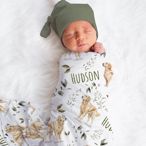 May include: A newborn baby wrapped in a white swaddle blanket and wearing a green knotted hat. The blanket features a pattern of golden retrievers, green leaves, and the name "HUDSON" in green lettering. The baby is lying on a fluffy white surface.