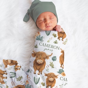 May include: A newborn baby wrapped in a white swaddle blanket and wearing a sage green knotted hat. The blanket features a repeating pattern of brown Highland cows, succulents, and the name "Cameron James" in green script.