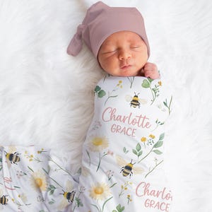 May include: A newborn baby wrapped in a white swaddle blanket and wearing a mauve knotted cap. The blanket features a floral and bee design with the name "Charlotte Grace" in pink script. The baby is lying on a white, fluffy surface.