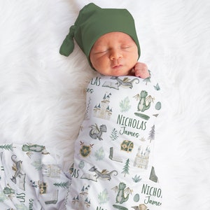 May include: A white swaddle blanket with a green, white, and gold dragon and castle pattern. The blanket is wrapped around a baby wearing a green hat. The blanket has the names "Nicholas James" printed on it.