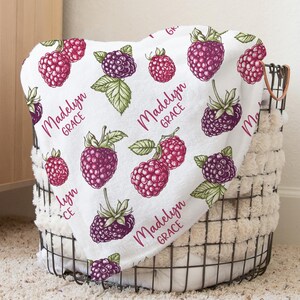 May include: A white fleece blanket with a repeating pattern of red and purple raspberries. The blanket has the name "Madelyn Grace" printed on it in a pink script font.