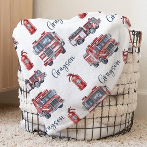 May include: A white fleece blanket featuring a repeating pattern of red fire trucks and red fire extinguishers, interspersed with the name Grayson written in a dark blue cursive script across the fabric surface.