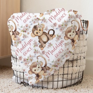 May include: A white baby blanket with a floral pattern and cute monkeys. The blanket is personalized with the name "Madelyn Grace" in pink lettering.