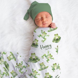 May include: A white swaddle blanket with a green frog pattern and the names "Henry Lucas" printed on it. A baby is swaddled in the blanket and is wearing a green hat.