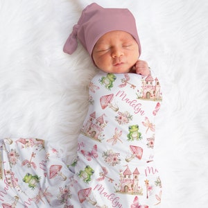 May include: A white swaddle blanket with a pink castle, flowers, butterflies, frogs, and dragonflies. The blanket has the name "Madelyn" written on it.