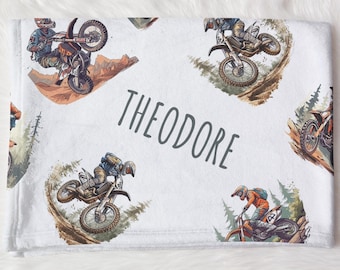 May include: White fleece blanket with a repeating pattern of motocross riders on dirt bikes. The blanket has the name "Theodore" printed in gray text.