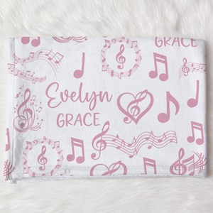 May include: A white baby blanket with a pink musical note pattern. The blanket features musical notes, treble clefs, and the names "Evelyn" and "Grace" in a soft pink font. The blanket is folded and displayed on a white, fluffy surface.
