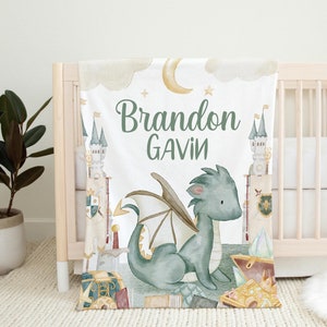 May include: A white baby blanket featuring a watercolor illustration of a friendly dragon, castle, and treasure chest. The blanket has the names "Brandon" and "Gavin" in green script. The design includes a moon, stars, and clouds.