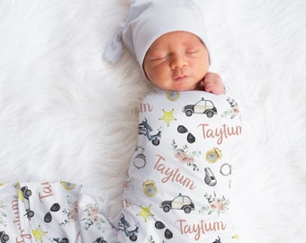 May include: A white swaddle blanket with a pink and blue police-themed pattern. The blanket features a repeating pattern of police cars, motorcycles, stars, sunglasses, and handcuffs. The name "Taytum" is written in pink script throughout the blanket.