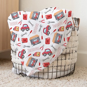 May include: A white baby blanket with a red car, wrench, and gas can pattern. The blanket has the name "Theodore" printed on it.