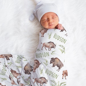 May include: A white swaddle blanket with a repeating pattern of brown bison and green leaves. The blanket is personalized with the name "HUDSON" in green text.