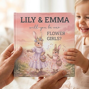 May include: A square book with a pink and orange sunset background featuring two illustrated rabbits wearing floral crowns and dresses, holding baskets. The text reads: LILY & EMMA will you be our FLOWER GIRLS? The cover art shows petals falling.