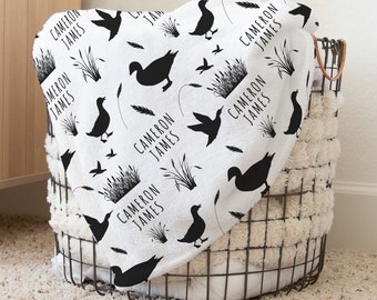 May include: A white baby blanket with a black and white pattern of ducks, reeds, and the name "Cameron James" repeated throughout.