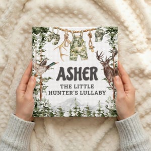 May include: A children's book titled "Asher The Little Hunter's Lullaby" with illustrations of hunting-themed items. The cover features a duck, deer, and camouflage overalls. The book is held by a person.