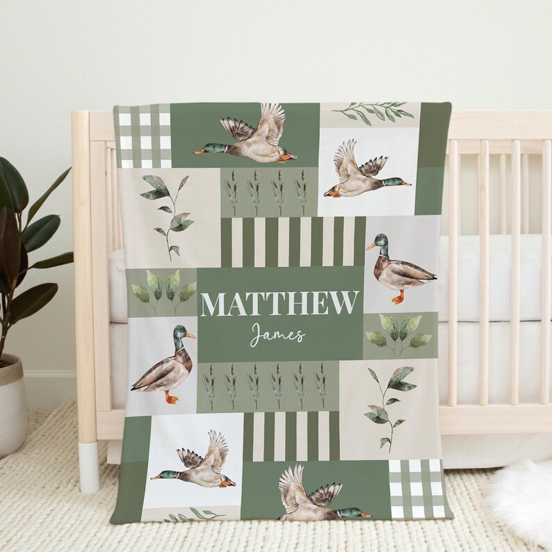 May include: A baby blanket with a patchwork design featuring ducks, foliage, and the name "MATTHEW James" in white. The blanket has a color palette of green, beige, and white, with a checkered pattern on the border.