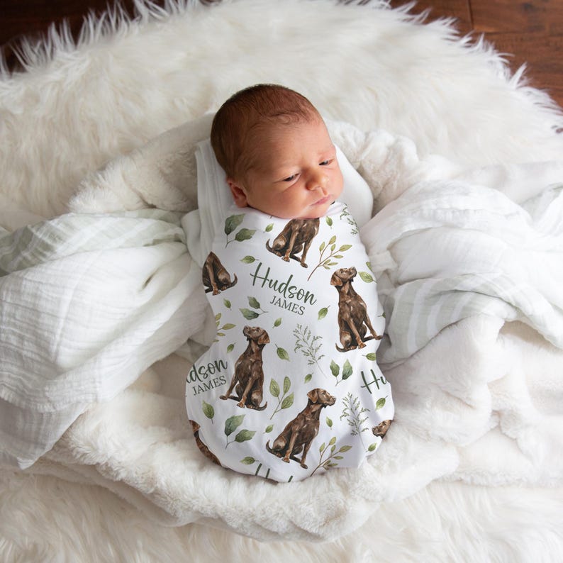 May include: A newborn baby wrapped in a white swaddle blanket with a repeating pattern of brown dogs and green leaves. The swaddle has the name "Hudson James" printed on it. The baby is lying on a fluffy white blanket.