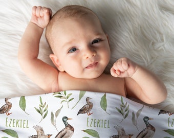 May include: A white baby blanket with a green and brown watercolor pattern of ducks and leaves. The blanket has the name "Ezekiel" printed on it.