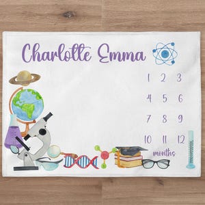 May include: A white baby blanket with a purple and blue science theme. It features a globe, a microscope, a beaker, a DNA strand, a molecule, a graduation cap, a book stack, a magnifying glass, safety goggles, a measuring cylinder, and a planet. The blanket has a space for the baby's name, "Charlotte Emma", and a chart to track their growth in months.