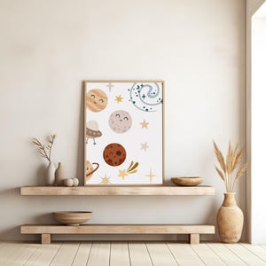 K&ouml;nnte beinhalten: Ein wei&szlig;er gerahmter Druck mit einer skurrilen Illustration von Planeten, Sternen und einer Galaxie. Die Planeten haben l&auml;chelnde Gesichter und sind in verschiedenen Farben, darunter Braun, Orange und Rosa. Die Sterne sind gelb und die Galaxie ist blau.