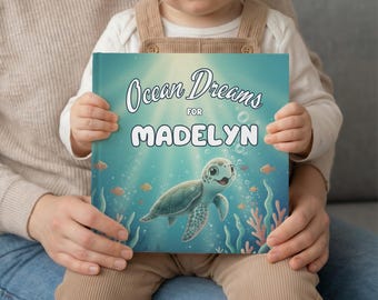 May include: A children's book titled "Ocean Dreams for Madelyn" is held by a child. The cover features a teal background with a cartoon sea turtle, fish, and coral. The title is in a script font.