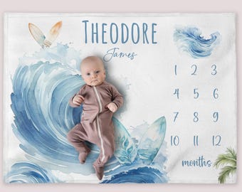 Puede incluir: Una manta de bebé blanca con un diseño de olas azules de acuarela y tablas de surf. La manta tiene una tabla de números para hacer un seguimiento del crecimiento de un bebé, con el texto "Theodore James" y "12 meses".
