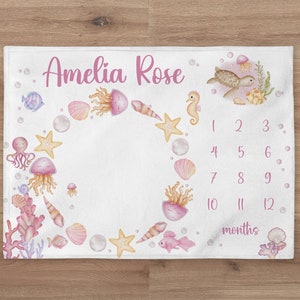 May include: White baby milestone blanket with a watercolor sea life design. Features the name "Amelia Rose" in pink script, surrounded by illustrations of jellyfish, starfish, and a seahorse. Includes a 1-12 months tracker.