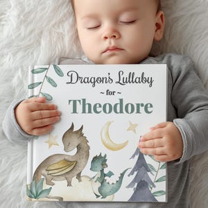 May include: A personalized children's book titled "Dragon's Lullaby" for Theodore. The cover features illustrations of dragons, a crescent moon, and stars in soft watercolor tones. The book is held by a baby.