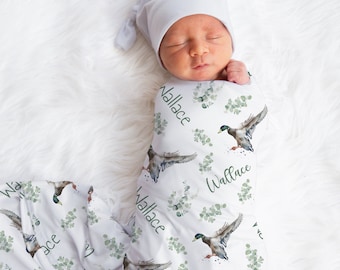 May include: A white swaddle blanket with a green eucalyptus and duck print. The blanket has the name "Wallace" repeated throughout the design. A baby is swaddled in the blanket and wearing a white hat.