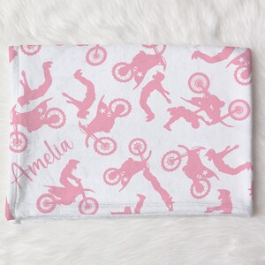 May include: Pink and white fleece blanket with a repeating pattern of pink motocross silhouettes. The blanket is personalized with the name "Amelia" in pink.
