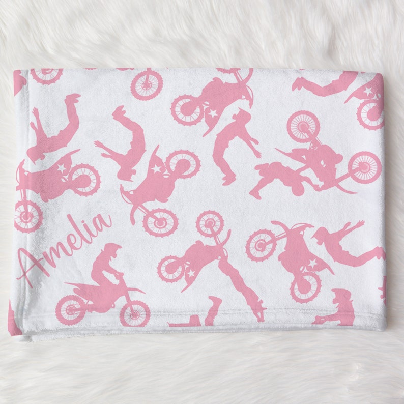 May include: Pink and white fleece blanket with a repeating pattern of pink motocross silhouettes. The blanket is personalized with the name "Amelia" in pink.