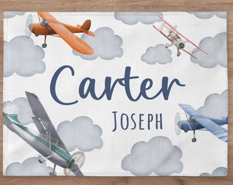 May include: A white blanket featuring watercolor illustrations of airplanes and clouds. The name "Carter" is written in navy blue script, with "JOSEPH" below. Orange, blue, and gray airplanes are scattered across the design.