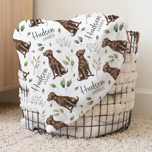 May include: A white blanket with a repeating pattern of brown dogs, green leaves, and the name "Hudson James" in black script. The blanket is folded over a wire basket filled with a fluffy, cream-colored material.