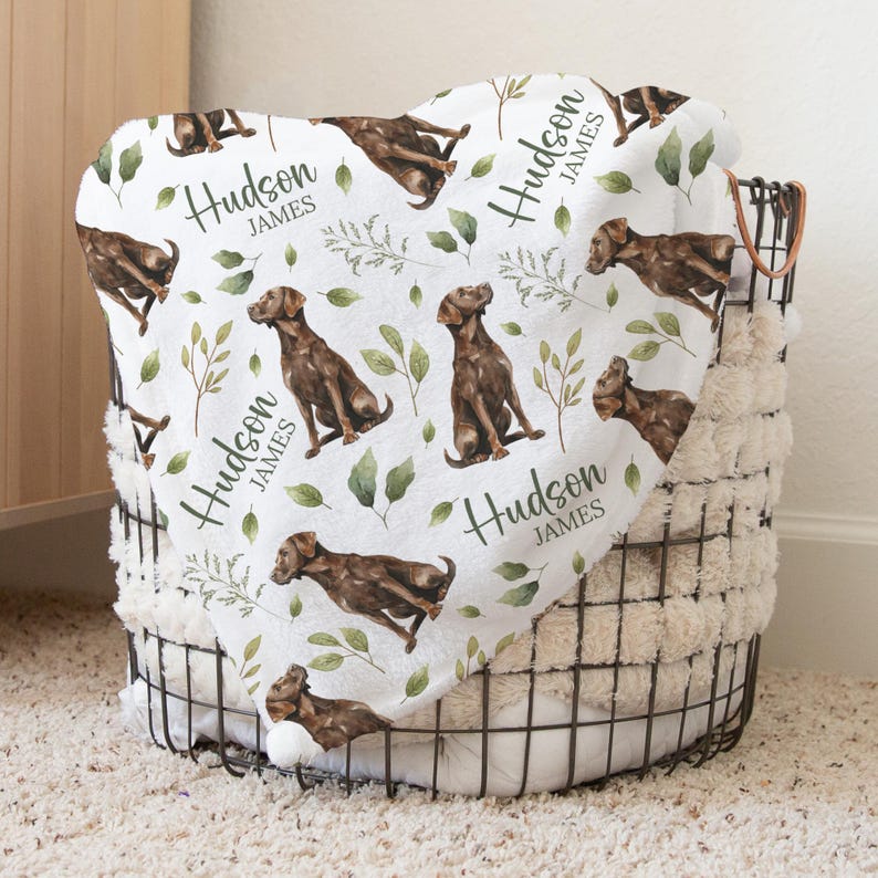 May include: A white blanket with a repeating pattern of brown dogs, green leaves, and the name "Hudson James" in black script. The blanket is folded over a wire basket filled with a fluffy, cream-colored material.