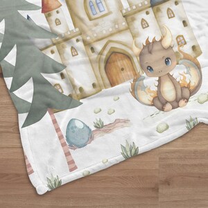 May include: A white fleece blanket with a watercolor illustration of a brown dragon with orange flames, a stone castle, and a blue egg. The dragon has large, blue eyes and is sitting on the ground.