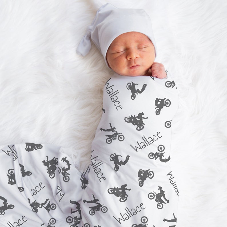K&ouml;nnte beinhalten: Ein Neugeborenes in einer wei&szlig;en Wickeldecke und mit einer grauen M&uuml;tze. Die Decke ist mit grauen Motorradsilhouetten und dem Namen "Wallace" gemustert. Das Baby liegt auf einer wei&szlig;en, flauschigen Oberfl&auml;che.