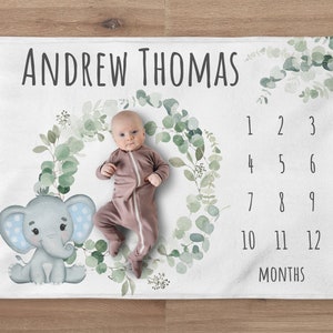 May include: A white baby blanket with a green eucalyptus wreath and a cute gray elephant with blue polka dots. The blanket has a number chart for tracking a baby's growth, with the text "ANDREW THOMAS" at the top.