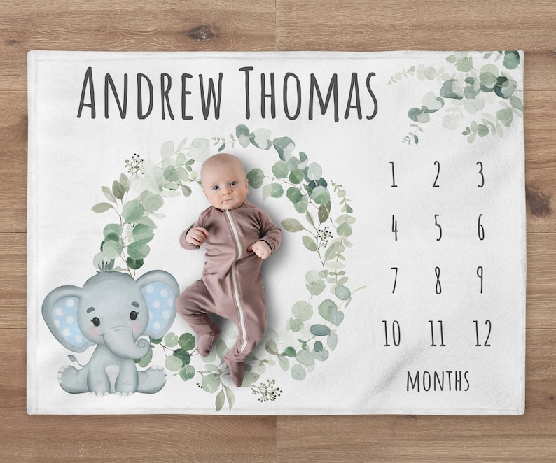May include: A white baby blanket with a green eucalyptus wreath and a cute gray elephant with blue polka dots. The blanket has a number chart for tracking a baby's growth, with the text "ANDREW THOMAS" at the top.