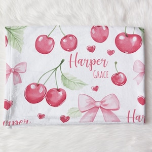 May include: White baby blanket with a pink and green cherry pattern. The blanket has the name "Harper Grace" written on it.