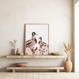 Peut inclure: Une impression d'art encadr&eacute;e repr&eacute;sentant deux canards orn&eacute;s de compositions florales. Les canards ont des t&ecirc;tes vertes, des corps bruns et des becs jaunes. L'&oelig;uvre est sur fond blanc et encadr&eacute;e dans un cadre en bois brun clair. Les compositions florales sont dans les tons de rose et de blanc.