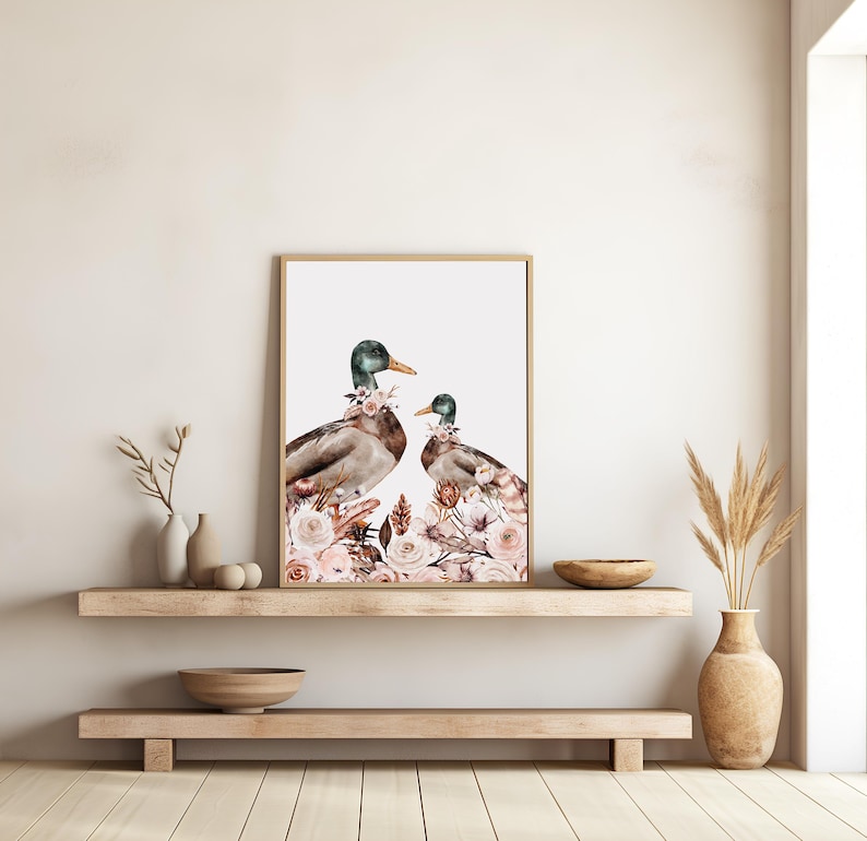 Peut inclure: Une impression d'art encadr&eacute;e repr&eacute;sentant deux canards orn&eacute;s de compositions florales. Les canards ont des t&ecirc;tes vertes, des corps bruns et des becs jaunes. L'&oelig;uvre est sur fond blanc et encadr&eacute;e dans un cadre en bois brun clair. Les compositions florales sont dans les tons de rose et de blanc.