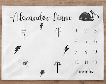 May include: White baby blanket with a black and white illustration of a hard hat, pliers, and lightning bolts. The blanket also has a number chart for tracking a baby's growth, with the names "Alexander Liam" at the top.