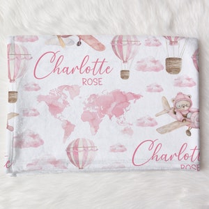 May include: Pink and white baby blanket featuring a world map, hot air balloons, airplanes, and clouds. The blanket has the name "Charlotte Rose" in cursive script. The design is a watercolor style.