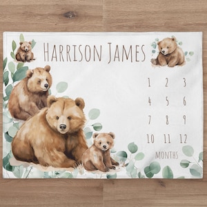 May include: A white baby milestone blanket with a watercolor design of brown bears and green leaves. The blanket has the name "Harrison James" printed on it and a chart for tracking the baby's growth in months.