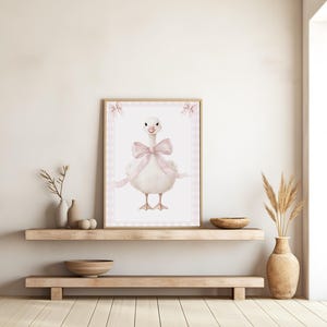 K&ouml;nnte beinhalten: Ein gerahmter Aquarelldruck mit einer bezaubernden wei&szlig;en Gans, die mit einer gro&szlig;en rosa Schleife geschm&uuml;ckt ist. Das Kunstwerk hat eine weiche, pastellfarbene Farbpalette und ist vor einem rosa Vichy-Muster angeordnet. Der Druck wird auf einem Holzregal ausgestellt.