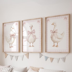 K&ouml;nnte beinhalten: Drei gerahmte Aquarelldrucke von G&auml;nsen, jede mit einer rosa Schleife. Die Drucke haben einen wei&szlig;en Hintergrund mit einem rosa Vichy-Rand und einem Holzrahmen. Unterhalb der Rahmen h&auml;ngt eine dekorative Wimpelkette.