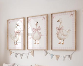 May include: Three framed watercolor prints of geese, each wearing a pink bow. The prints are set against a white background with a pink gingham border and a wooden frame. A decorative banner of small flags hangs below.